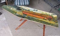 Original oil painting on ironing board with scenic rural landscape including covered bridge and farm buildings, placed on wooden stand.