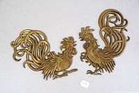 Pair of vintage gold-tone metal roosters with scrollwork tails laid on flat white surface, showing full front views.