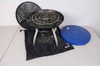 Full view of the Coleman portable propane grill with cooking grate, blue carrying case, and black fabric bag on surface.