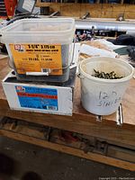 Image showing a plastic toolbox filled with 1-1/4 inch coarse thread drywall screws, a box of coil roofing nails, and a white bucket marked 12D sinker containing various nails.