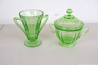 Two pieces of green uranium glass: a sugar bowl with lid and a two-handled cup or vase, shown side by side on a white surface.
