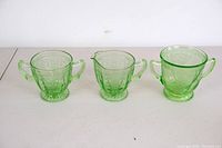 Set of three green uranium glass vessels: one sugar bowl and two creamers, on a flat surface, showing size and shape differences.