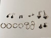 Full view of 8 pairs of silver earrings arranged on a white surface showing variety of styles including hoops, drop earrings with black stones, studs, and triangular dangle earrings.