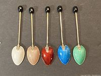 Five sterling silver enamelled coffee spoons arranged side by side, showing the colorful enamel on the top side of the spoon bowls in white, beige, red, blue, and green.