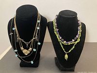 Full view of multiple necklaces on two busts showing layered gold and silver chains with pale blue beads and flower pendant, plus double strand green and purple stone bead necklaces.