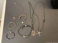 Full view of seven pieces of silver jewelry including bracelets and necklaces on a dark surface.