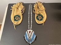 Pair of tan leather moccasins with circular beadwork and fringed edges, alongside the beaded necklace laid flat on a dark surface.