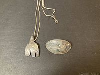 Photo showing the animal-shaped Haida silver pendant on a silver chain and the oval Haida pendant/brooch placed side by side on a dark background.