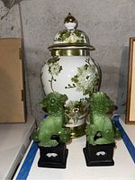 Photo showing the ceramic ginger jar decorated with green floral vines and gold trim, alongside two green Lucite lion figurines on black bases.