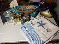 View of the lot items including the decorative bowls, brass mortar and pestle, glass jar, perfume bottle, wall hanging, and tablecloth, arranged on a table.