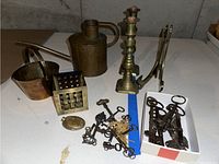 Photo showing copper watering can, brass pail, two brass candlesticks, brass box, fork-shaped brass candle holders, and vintage keys on table surface.