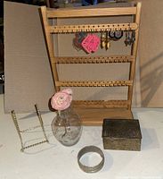 Wooden earring stand with five pairs of hanging earrings, glass vase with faux flower, metal easel, trinket box and cuff bracelet displayed together.