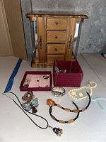 Photo showing wood jewelry box with three drawers and glass side panels, yarn trinket box, various pins, Geneva bracelet watch, and several necklaces including purple stone pendant and carved wood bead necklace.