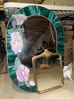 Oval mirror with stained glass floral frame shown with smaller gold framed mirror in front on basement floor.
