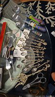 Full lot of assorted serving utensils including spoons, forks, small servers and scissors laid out on a glass table, showing colorful large handles and ornate small spoons.