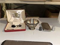 Photo showing the full lot: cufflinks in case, silver bowl, lighter cover, and thimble arranged on a table.