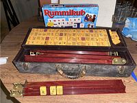 Open carrying case with mahjong tiles arranged in rows and held by brass and bakelite trays, with the Rummikub game box visible behind.