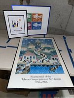 Photo showing two framed artworks; one large poster with a colorful community procession and textual commemoration, and one smaller colorful fish-themed signed print.