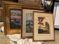 Three framed landscape paintings displayed together showing different scenes including a coastal village, rural house, and textured landscape.