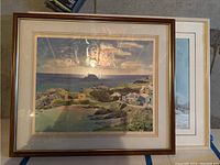 Two framed wall art prints with coastal themes, one over the other on a concrete floor.