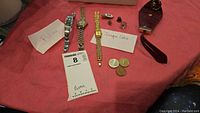 Overview of all items including three watches labeled 'Pulsar', 'Jacquito Paris', and one unidentified (likely Anne Klein), coins, tie clip, key holder, and small accessories on a pink cloth surface.