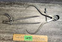 Two metal calipers placed on a concrete surface with a wooden ruler for scale showing approximately 6 inches length. The calipers have pointed ends and mechanical adjustment.