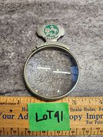 Full view of circular magnifying glass showing metal frame with inscriptions and enamel emblem on top.