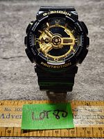 Front view of Casio G-Shock watch face showing black and gold design, 'G-SHOCK' and 'PROTECTION' in gold lettering on bezel, placed on gray textured surface with measuring ruler below.