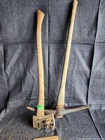 Two axes standing upright on a black fabric background, showing the splitting axe on left and pick axe on right, both with wooden handles and metal heads.
