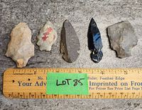 Five stone arrowheads arranged side by side above a wooden ruler showing sizes ranging from about 1.5 to 4 inches. Arrowheads made from different colored stones.
