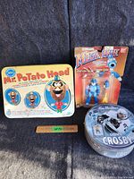 Photo showing all three items: Mr. Potato Head metal box, Mega Man figure in original packaging, and Tim Hortons puzzle tin.