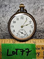 Front view of the small vintage pocket watch showing white dial with black Arabic numerals, gold-tone hour and minute hands and a sub-second dial at 6 o'clock. Size shown against ruler indicating about 1.5 inches diameter.