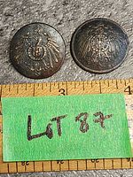 Two brass WW2 German military uniform buttons with eagle crest design placed on stone surface with a measuring tape beneath them showing roughly 3.25-inch total width for both buttons together.