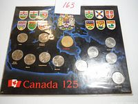 Full view of the Canada 125th Anniversary Provincial Coin Set displayed on a black board with a blue map background, featuring provincial shields and the Canadian coat of arms, showing multiple silver-colored coins in place representing provinces and territories.