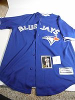 Full view of blue Toronto Blue Jays Majestic jersey with buttons, showing logo and text, with rookie card placed on jersey.