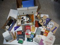 Wide view of the lot showing boxes, papers, envelopes, photo collage book, calculator, playing cards, and various small office supplies arranged on the floor.