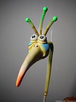Close-up photo of the metal sculpture's head showing the long yellow-orange beak, bead eyes, and green-tipped coiled antennae.