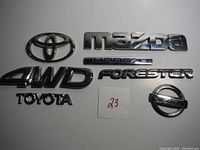 Image showing multiple vintage vehicle emblems including Toyota oval logo, Mazda, Subaru Impreza, Forester, Nissan, 4WD, and Toyota text badges arranged on flat surface.