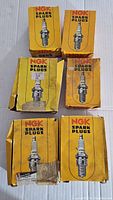 Six vintage NGK spark plug boxes, yellow-orange with spark plug image, some wear.