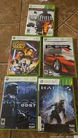 Photo of five Xbox 360 game cases arranged on tile floor showing the cover art for Battlefield Bad Company 2, Star Wars The Clone Wars, PGR 4, Halo 3: ODST, and Halo 4.