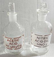 Front view of two clear glass Pyrex acid jars with white chemical labels, one labeled 'Acetic Acid HC2H3O2', the other 'Nitric Acid Dil. HNO3'. Both jars have stoppers and are about 6 inches tall.