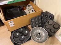 Photo showing four muffin pans, Bundt pan, pizza pan, wooden rolling pin, and two fabric oven mitts inside a wooden box, and some displayed outside on floor.