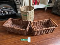 Overall view of two rectangular baskets with wood handles placed side-by-side and one taller round basket in the background on wooden floor in front of fireplace.