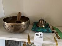 Wide view showing the brass singing bowl with wooden mallet inside, and the brass bell on a blue cushion with gold tassels, all placed on a white shelf.