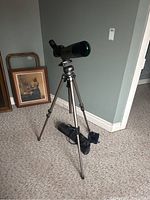 Celestron spotting scope mounted on tripod in room corner with paintings leaning on wall.