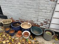 Photo showing a collection of 11 ceramic clay pots arranged outdoors near a brick wall on leaf-covered ground.