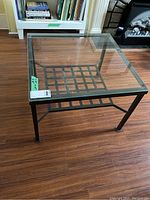 Full view of square coffee table with clear glass top and black metal frame base with grid shelf underneath on wooden flooring.
