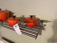 Two small flame orange saucepan-style pots with lids and long wooden handles, sitting on metal rack shelf.