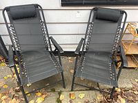 Two black folding lounge chairs side by side outdoors on concrete ground near white siding wall. Chairs have metal frames, black mesh fabric seats and backs with lace-up sides, padded black headrests, and armrests.