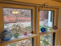 Photo showing all 8 handblown glass ornaments arranged on a narrow window shelf, highlighting the different sizes and colors including blue, green, pink, amber, and purple spheres.
